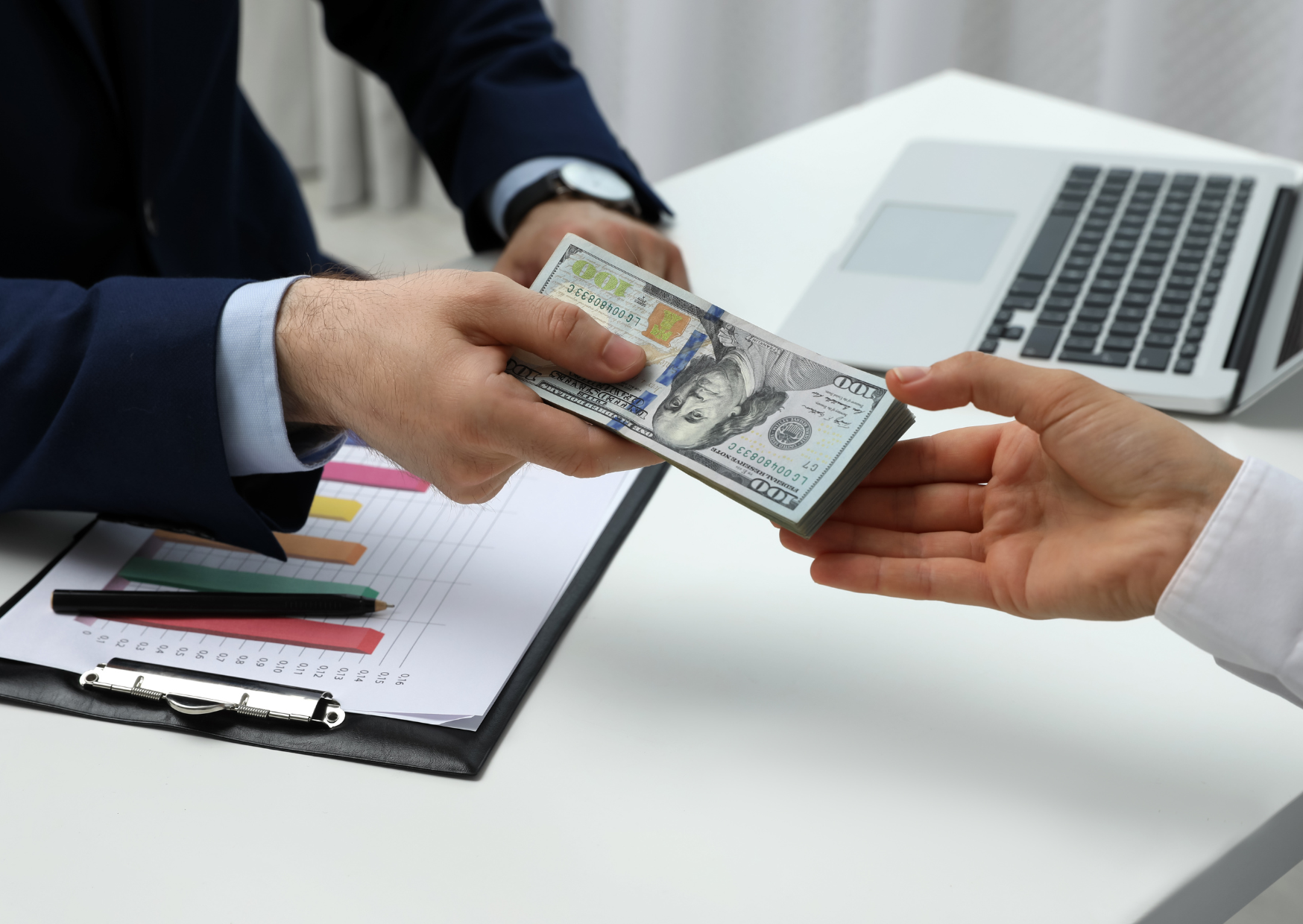 Two people exchanging a stack of U.S. hundred-dollar bills at a desk with financial charts and a laptop nearby.
