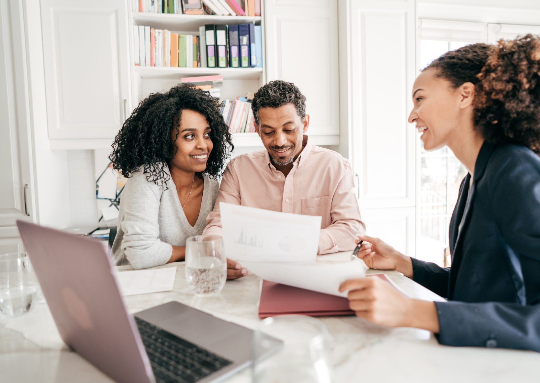 Couple reviews financial documents with smiling advisor at kitchen table, discussing real estate loan options.